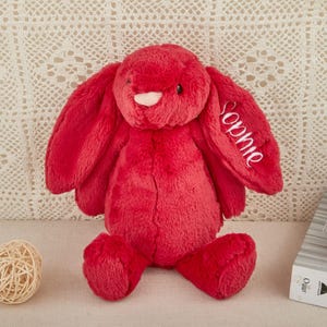 May include: A plush, red bunny rabbit with long floppy ears. The name "Sophie" is embroidered in white script on one ear. The bunny is sitting upright, with its arms and legs extended. A decorative ball and a string of pearls are in the foreground.