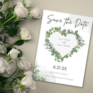May include: A white save-the-date card with a heart-shaped wreath of green leaves and the names "Oliver and Juliana." The card also includes the date "6.21.25" and the text "Brix Napa Valley, Formal Invitation to Follow." White roses are in the background.