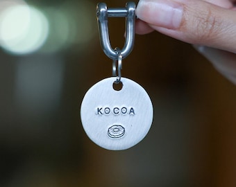 May include: A silver-colored keychain tag with the word "KO COA" and a donut graphic. The tag is round and attached to a metal clasp. The keychain is held up by a person's hand.