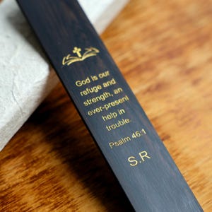 Könnte beinhalten: Dunkles Holzlesezeichen mit goldfarbenem Schriftzug. Der Text lautet: „Gott ist unsere Zuflucht und Stärke, eine immer gegenwärtige Hilfe in der Not. Psalm 46:1 S.R.“ Oben befindet sich ein goldenes Kreuz- und offenes Buch-Design.