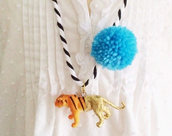 May include: A white shirt with a black and white rope necklace with a blue pom pom and a gold tiger pendant.