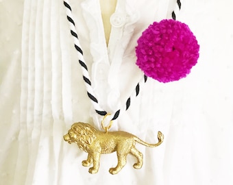 May include: A gold lion pendant hangs from a black and white striped cord necklace with a large pink pom-pom at the end.