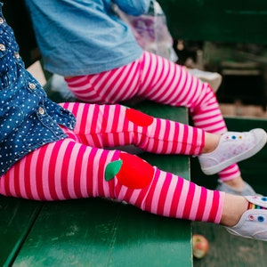 May include: Two children wearing pink and white striped leggings with apple patches on the knees. The leggings are paired with white sneakers. The children are sitting on a green bench.