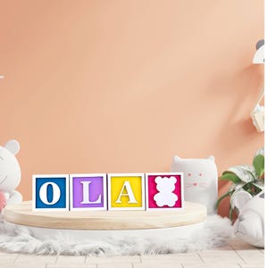 May include: Wooden blocks spelling "OLA" in blue, purple, yellow, and pink, with a white bear outline on the last block. The blocks sit on a round, light-coloured platform with a fluffy white rug.