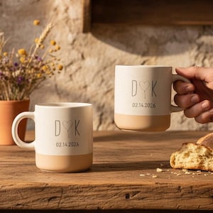 Könnte beinhalten: Zwei cremefarbene Keramikbecher mit beigefarbenem Boden, jeweils mit den Initialen "D♡K" und dem Datum "02.14.2026". Ein Becher wird von einer Person gehalten. Die Becher stehen auf einem Holztisch mit Brot und einem kleinen Terrakottatopf mit Trockenblumen.