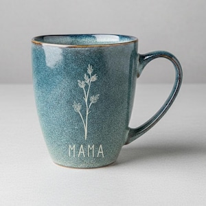Puede incluir: Una taza de cer&aacute;mica color turquesa con un dise&ntilde;o floral blanco y la palabra "MAMA" impresa en la parte delantera. La taza tiene forma redondeada, un asa y un borde dorado. La taza mide aproximadamente 10 cm de alto.