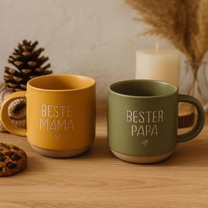 Può includere: Due tazze in ceramica, una giallo senape e una verde oliva, con le parole "BESTE MAMA" e "BESTER PAPA" in bianco. Ogni tazza ha un simbolo a cuore sotto il testo. Le tazze sono su una superficie di legno.