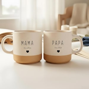 Pode incluir: Duas canecas de cerâmica com a parte superior creme e a base bege. Uma caneca diz "MAMA" com "Est.2026" e um coração. A outra caneca diz "PAPA" com "Est.2026" e um coração. Ambas as canecas têm uma alça curva.