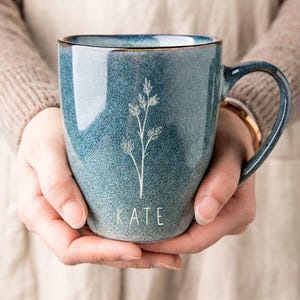 Puede incluir: Una taza de cer&aacute;mica azul con asa, con un dise&ntilde;o floral blanco y el nombre "KATE" en la parte inferior. La taza tiene un efecto degradado, pasando de un azul m&aacute;s oscuro en la parte superior a un azul m&aacute;s claro en la parte inferior. El borde tiene un ribete marr&oacute;n.