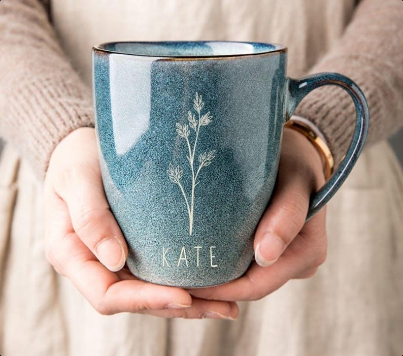 Puede incluir: Una taza de cer&aacute;mica azul con asa, con un dise&ntilde;o floral blanco y el nombre "KATE" en la parte inferior. La taza tiene un efecto degradado, pasando de un azul m&aacute;s oscuro en la parte superior a un azul m&aacute;s claro en la parte inferior. El borde tiene un ribete marr&oacute;n.