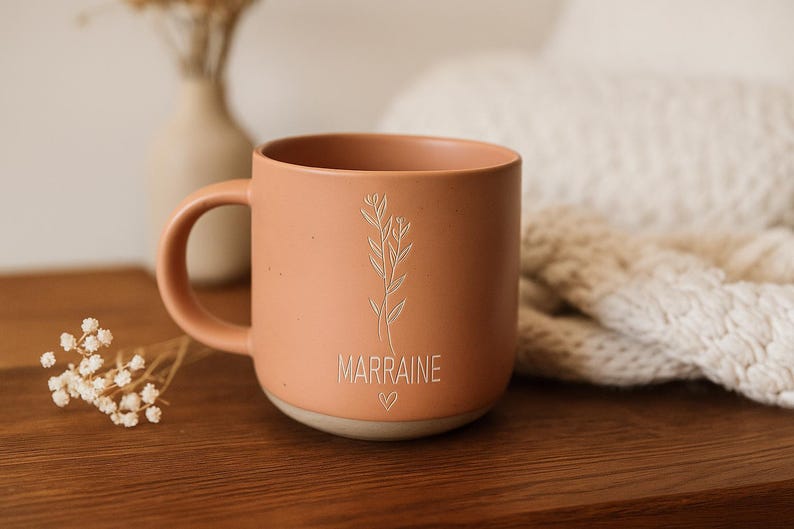 May include: A peach-colored ceramic mug with a handle, featuring a floral design and the word "MARRAINE" in a delicate font. A small heart is below the word. The mug sits on a wooden surface.