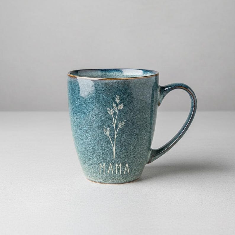 Puede incluir: Una taza de cer&aacute;mica color turquesa con un dise&ntilde;o floral blanco y la palabra "MAMA" impresa en la parte delantera. La taza tiene forma redondeada, un asa y un borde dorado. La taza mide aproximadamente 10 cm de alto.