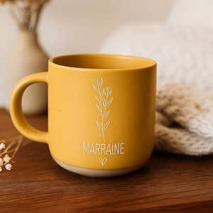 May include: Mustard yellow ceramic mug with a handle, featuring a floral design and the word "MARRAINE" with a heart symbol. The mug sits on a wooden surface, with a vase and dried flowers in the background.