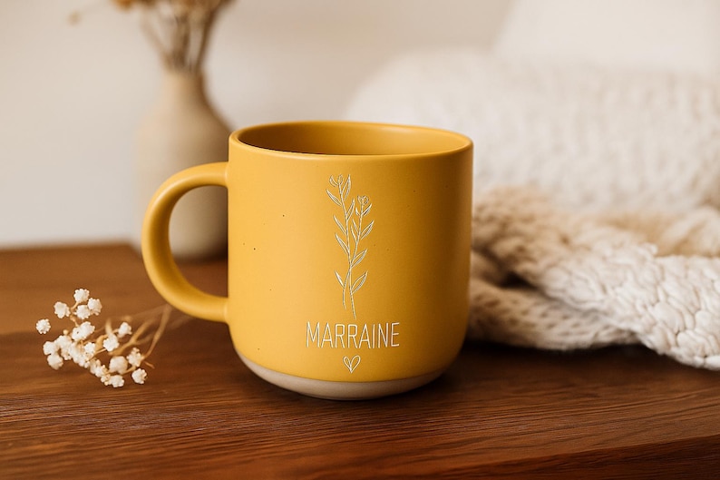 May include: Mustard yellow ceramic mug with a handle, featuring a floral design and the word "MARRAINE" with a heart symbol. The mug sits on a wooden surface, with a vase and dried flowers in the background.