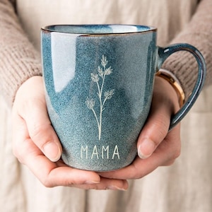 Puede incluir: Una taza de cer&aacute;mica azul con asa, con un dise&ntilde;o floral blanco y la palabra "MAMA". La taza tiene un acabado moteado y un borde marr&oacute;n. La taza se sostiene con ambas manos.
