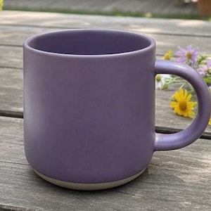 May include: A solid purple ceramic mug with a rounded base and a curved handle. The mug has a matte finish and a light beige base. The mug is sitting on a wooden surface, with yellow and purple flowers in the background.