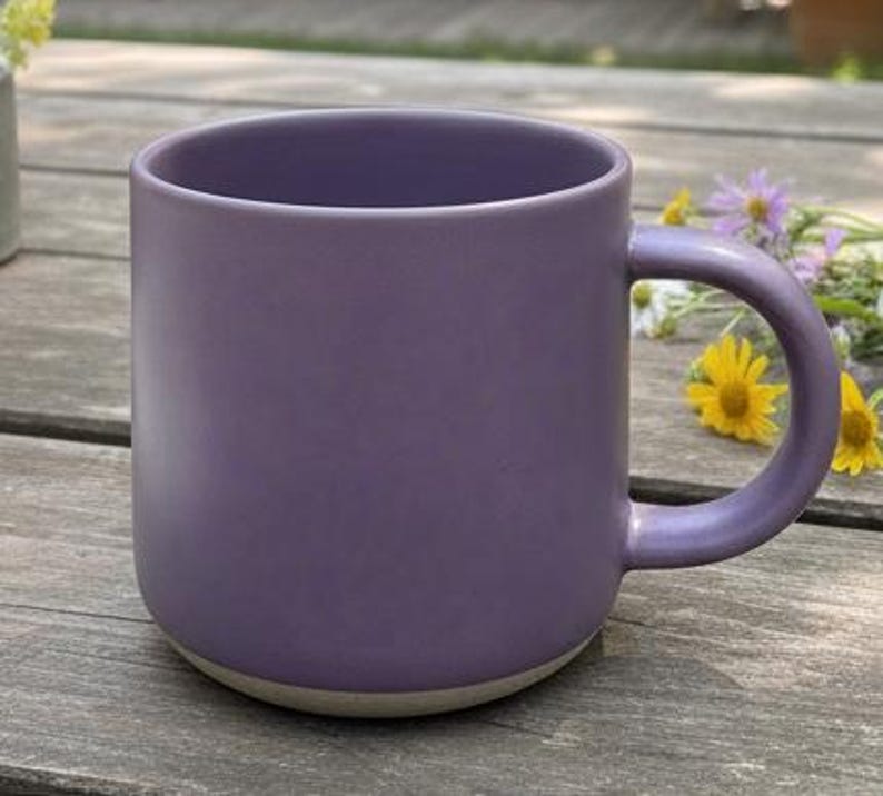 May include: A solid purple ceramic mug with a rounded base and a curved handle. The mug has a matte finish and a light beige base. The mug is sitting on a wooden surface, with yellow and purple flowers in the background.