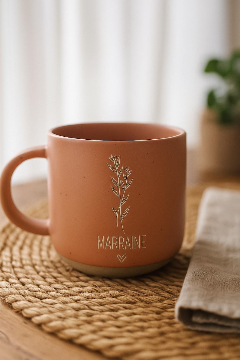 May include: Terracotta-colored ceramic mug with a handle, featuring a floral design and the word "MARRAINE" with a heart symbol. The mug sits on a woven placemat, with a neutral-colored napkin nearby. A plant is visible in the background.