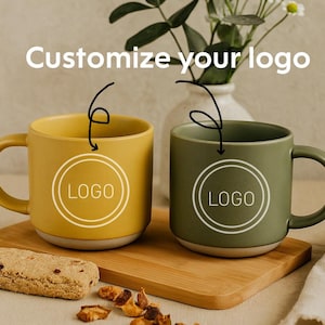 Może przedstawiać: Dwa ceramiczne kubki, jeden musztardowy i jeden oliwkowy, z białym wzorem "LOGO", stoją na drewnianej desce. Tekst "Customize your logo" znajduje się nad kubkami. Ciastko i orzechy są na pierwszym planie.