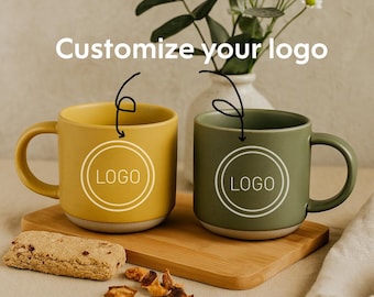 May include: Two ceramic mugs, one mustard yellow and one olive green, with a white "LOGO" design, sit on a wooden board. The text "Customize your logo" is above the mugs. A cookie and nuts are in the foreground.