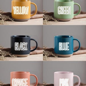 May include: A collection of ceramic mugs in various colors: yellow, green, black, blue, orange, and pink. Each mug has a contrasting base and a white label with the color name. The mugs are displayed on a wooden surface.