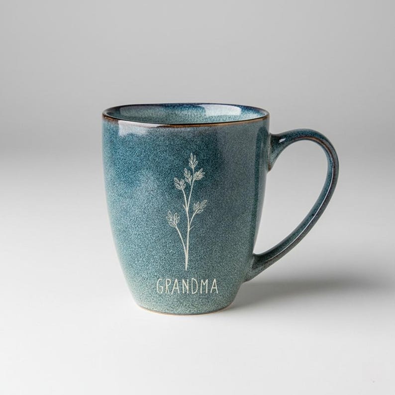 Puede incluir: Una taza de cer&aacute;mica color turquesa con borde marr&oacute;n y asa curva. La taza presenta un dise&ntilde;o floral blanco y la palabra "GRANDMA". La taza tiene una textura moteada y un efecto de color degradado.