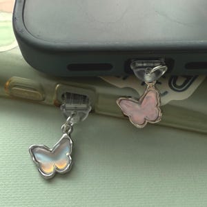 May include: Two butterfly phone charms. One charm has a pearlescent white and yellow butterfly with a silver-colored border. The other charm has a pink butterfly with a silver-colored border. Both charms are attached to a phone.