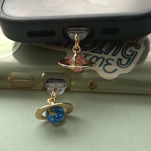 May include: Two gold-colored planet charms attached to a phone. One charm features a pink planet with a gold ring, the other a blue planet with a gold ring. The phone is black and has a clear case.