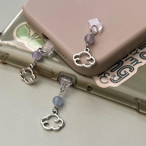 May include: Two phone charms with silver cloud charms and purple and blue beads are attached to a phone case. The phone case is a light tan color and is resting on top of a clear phone case with a sticker.