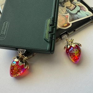 May include: Two phone cases with strawberry charms. One case is dark green with a textured surface. The other case is black with a sticker. The strawberry charms are red, gold, and iridescent.