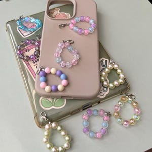 May include: A collection of colorful beaded phone charms in various pastel colors, including pink, blue, purple, and white. The charms are circular and attached to silver clasps. Some charms are on a pink phone case.