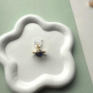 May include: A gold-colored planet charm earring with a blue planet and a crescent moon design. The earring is attached to a clear earring clip. The earring is displayed on a white, cloud-shaped tray.