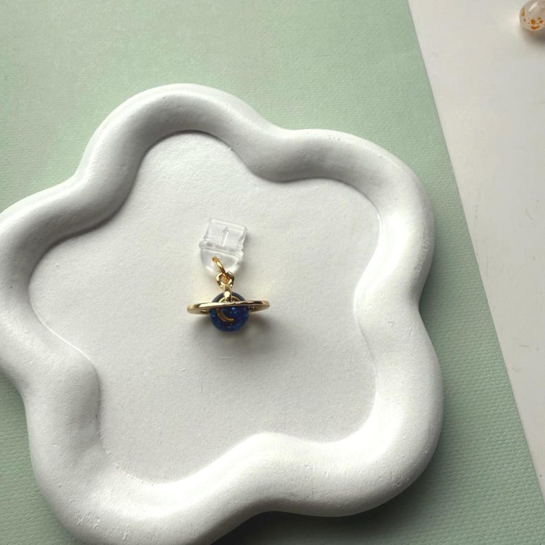 May include: A gold-colored planet charm earring with a blue planet and a crescent moon design. The earring is attached to a clear earring clip. The earring is displayed on a white, cloud-shaped tray.