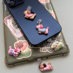 May include: A collection of colorful dog-shaped phone charms. The charms are in various poses and outfits, including pink, blue, and patterned clothing. The charms are attached to a phone case and phone.