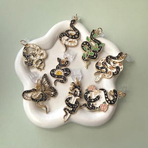 May include: A collection of gold-toned earrings with black and white snake designs, a butterfly, and floral accents. The earrings are displayed on a white, flower-shaped tray against a light green background.