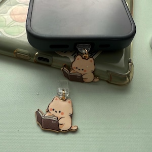 May include: Two beige cat charms, each reading a book, are attached to a phone case. The charms have gold-colored accents and are hanging from the phone case. The phone case is clear with a green tint.