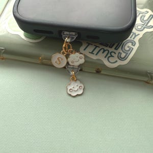 May include: A gold-colored phone charm with white cloud and letter "S" charms. The cloud charms have smiling faces. The phone case is dark gray.