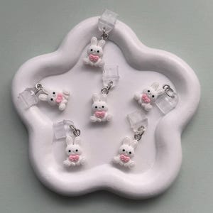 May include: White bunny earrings with pink bow ties are displayed on a white, cloud-shaped tray. The earrings have silver-colored hooks and clear earring backs. The earrings are a cute, whimsical accessory.
