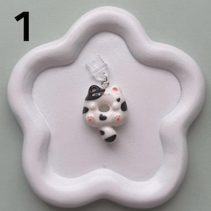 May include: A white and black cat-shaped earring with a donut-like body, black spots, and pink paw pads. The earring is attached to a clear earring post and is set on a white, cloud-shaped tray. The number "1" is in the upper left corner.
