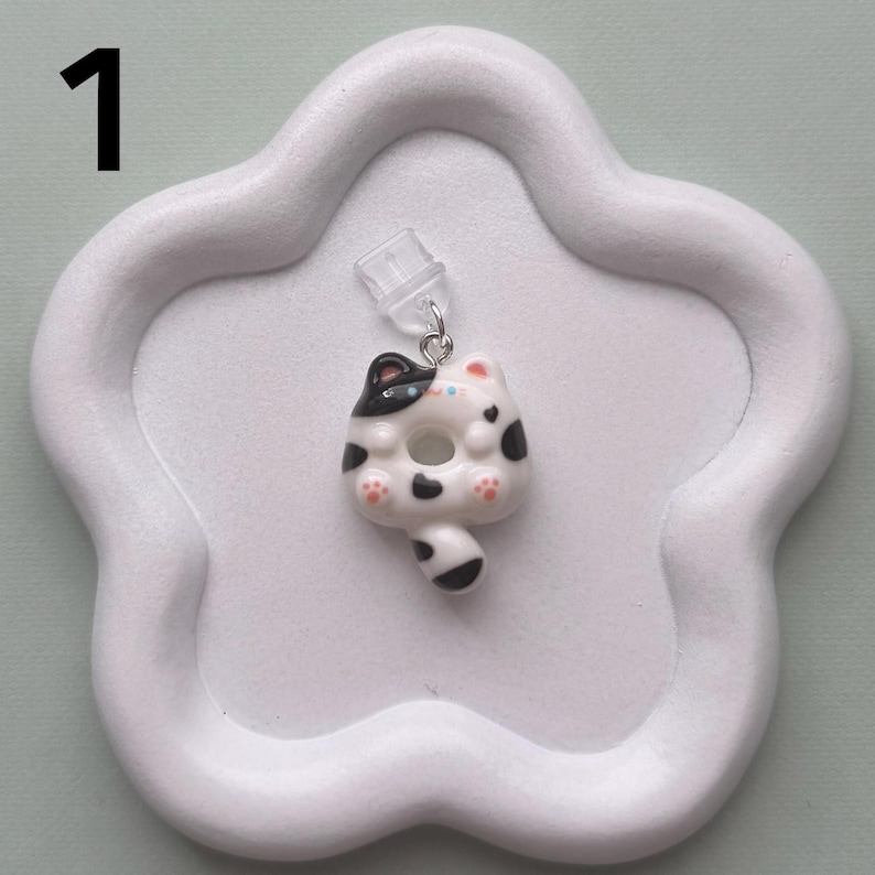 May include: A white and black cat-shaped earring with a donut-like body, black spots, and pink paw pads. The earring is attached to a clear earring post and is set on a white, cloud-shaped tray. The number "1" is in the upper left corner.