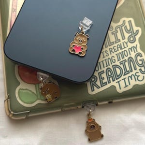 May include: Brown bear phone charms with gold accents. One bear holds a pink heart, another a drink, and the third wears a crown. The charms are attached to a phone and phone case, with a green background.