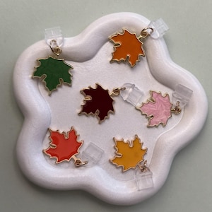 May include: A collection of colorful maple leaf earrings in shades of green, orange, red, yellow, pink, and burgundy, each with a gold-colored outline. The earrings are displayed on a white, cloud-shaped tray.