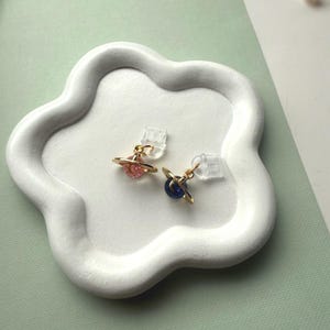 May include: Two planet-shaped stud earrings with gold-colored rings and colorful centers, one pink and one blue. The earrings are displayed on a white, cloud-shaped tray. The background is a soft green.