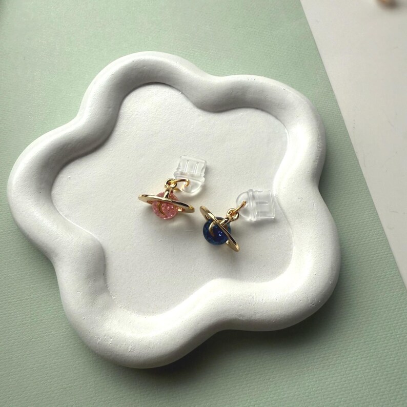 May include: Two planet-shaped stud earrings with gold-colored rings and colorful centers, one pink and one blue. The earrings are displayed on a white, cloud-shaped tray. The background is a soft green.