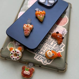May include: A collection of colorful phone charms in the shape of food items, including a donut, croissant, fish, and pastries. The charms are attached to a phone case and a blue phone.