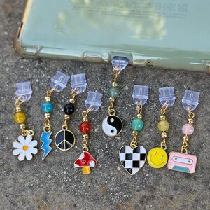 May include: A collection of colorful phone charms with various designs, including a daisy, lightning bolt, peace sign, mushroom, yin-yang symbol, checkered heart, smiley face, and cassette tape. Each charm has a gold-colored chain and colorful beads.