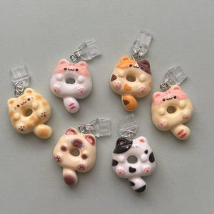 May include: A collection of six cat-shaped donut charms in various colors, including yellow, orange, pink, and white. Each charm has a clear clip and silver-colored metal ring attached. The charms are designed with cute details, such as paw prints and facial features.
