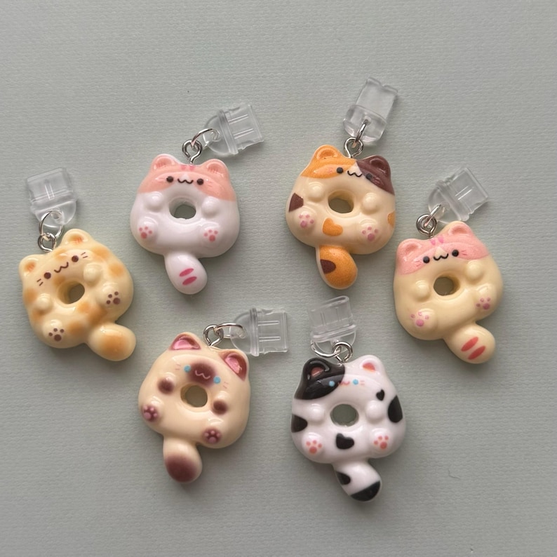 May include: A collection of six cat-shaped donut charms in various colors, including yellow, orange, pink, and white. Each charm has a clear clip and silver-colored metal ring attached. The charms are designed with cute details, such as paw prints and facial features.