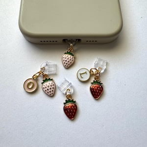 May include: A collection of phone charms featuring strawberry and initial designs. The charms have gold-colored metal accents and are attached to clear port plugs. The strawberries are pink and red with green tops, and the initial charms have white letters.