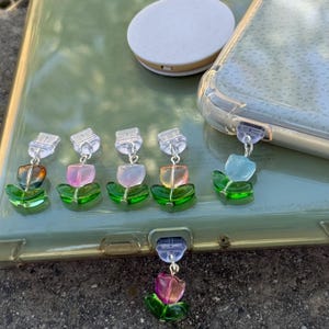 May include: A collection of colorful flower-shaped phone charms. Each charm features a translucent flower with green leaves and a silver-colored chain. The flowers are in various colors, including orange, pink, and blue. A white phone grip and a phone case are also visible.